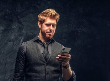 Formal giyim bir smartphone stüdyoda bir karanlık dokulu duvara tutan yakışıklı Kızıl saçlı adam