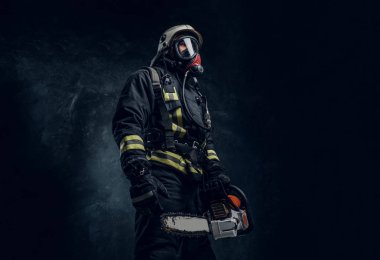 Kask ve oksijen maskesi bir chainsaw tutan bir itfaiyeci portresi. Studio fotoğraf bir karanlık dokulu duvara