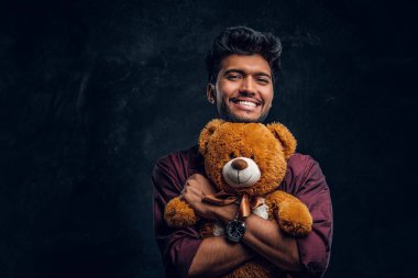 Genç Hintli adam şık gömlek gülümseyen bir oyuncak ayı ve yana doğru seyir hugs.