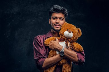 Yakışıklı genç Hintli şık gömlek hugs oyuncak ayı ve a fotoğraf makinesi vasıl arıyor.
