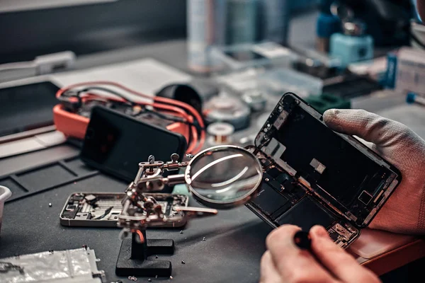 Serviceman utiliza lupa y destornillador para reparar el teléfono inteligente dañado en el taller . —  Fotos de Stock