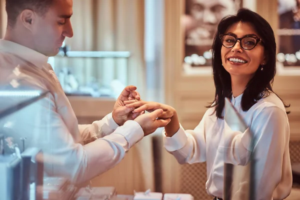 Elegantly dressed man trying on the wedding ring on the hand of his girlfriend in a jewelry store