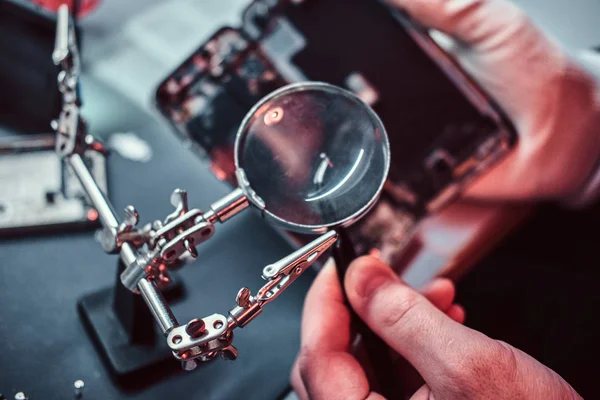 Repairman uses magnifier and tweezers to repair damaged smartphone. — Stock Photo, Image