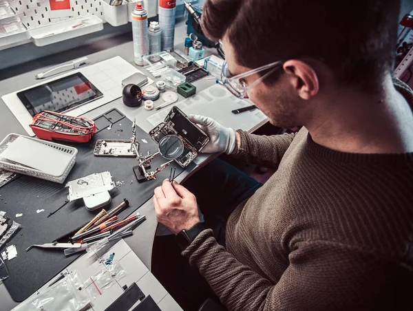 Служащий использует увеличитель и пинцет для ремонта поврежденного смартфона в мастерской . — стоковое фото