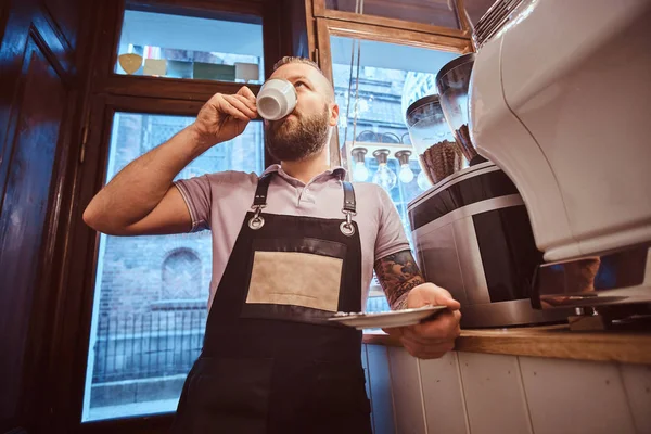 Barista guapo en delantal bebiendo café durante la pausa para el almuerzo apoyado en un mostrador en la cafetería — Foto de Stock