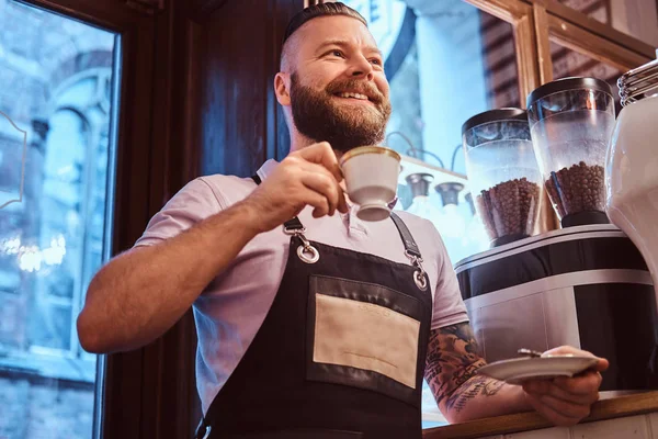 Happy barista em avental segurando uma xícara e um pires, bebendo café durante a pausa para o almoço apoiando-se em um balcão na cafeteria . — Fotografia de Stock