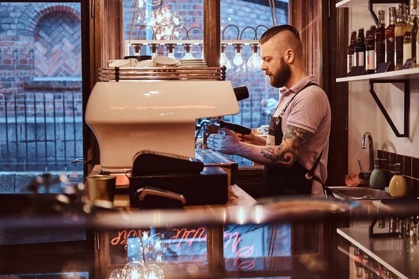 Τατουάζ barista με στιλάτα γένια και το χτένισμα που εργάζεται πάνω σε μια μηχανή του καφέ σε μια καφετέρια ή εστιατόριο — Φωτογραφία Αρχείου