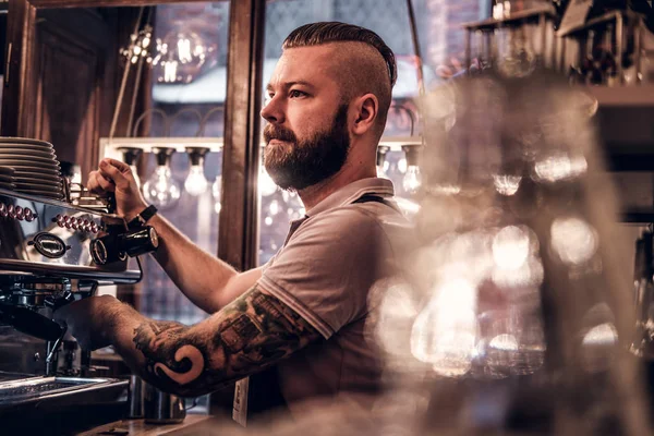 Tattooed barista with stylish beard and hairstyle working on a coffee machine in a coffee shop or restaurant — Stock Photo, Image