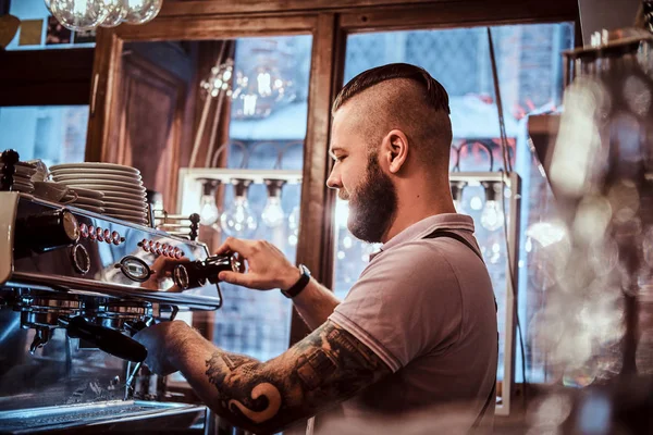 Τατουάζ barista με στιλάτα γένια και το χτένισμα που εργάζεται πάνω σε μια μηχανή του καφέ σε μια καφετέρια ή εστιατόριο — Φωτογραφία Αρχείου
