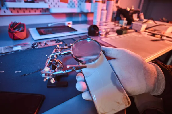 Elektroniker hält ein modernes Smartphone mit kaputtem Körper in der Hand, untersucht den Schaden sorgfältig mit einer Lupe. Beleuchtung mit rotem und blauem Licht — Stockfoto