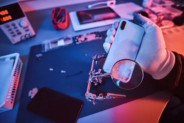 Elektroniker hält ein modernes Smartphone mit kaputtem Körper in der Hand, untersucht den Schaden sorgfältig mit einer Lupe. Beleuchtung mit rotem und blauem Licht — Stockfoto