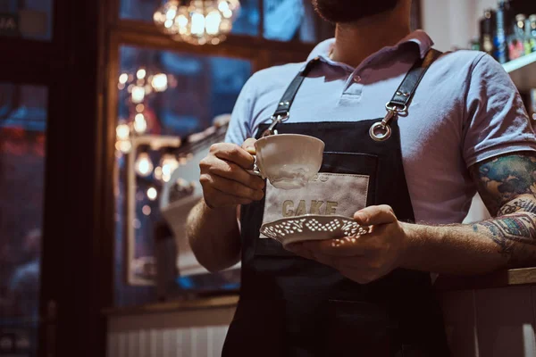 Barista con delantal sosteniendo una taza y un platillo, tomando café durante la pausa para el almuerzo apoyado en un mostrador en la cafetería . — Foto de Stock