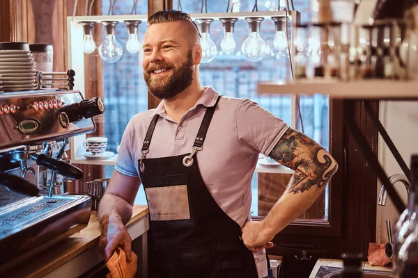 Barista with stylish beard and hairstyle wearing apron smiling and looking sideways while leaning on a counter in the cafe or restaurant