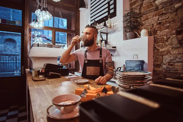 Barista en delantal tomando café durante la pausa para el almuerzo de pie detrás del mostrador en la cafetería —  Fotos de Stock