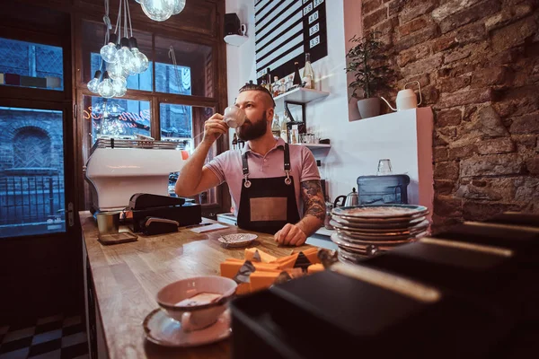 Barista en delantal tomando café durante la pausa para el almuerzo de pie detrás del mostrador en la cafetería — Foto de Stock