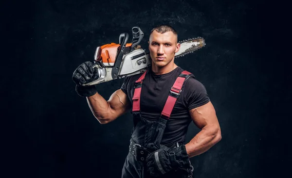 Brutal logger posing with a chainsaw on his shoulder and looking at a camera with a confident look — Stock Photo, Image