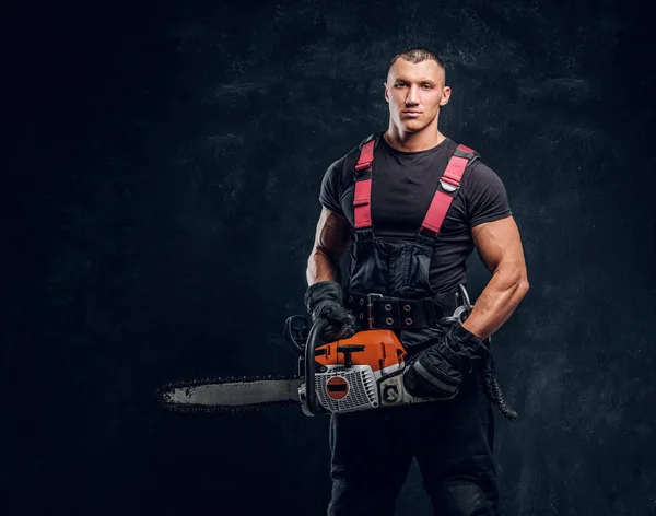 Young lumberjack wearing protective clothes posing with a chainsaw in a dark studio — Stock Photo, Image