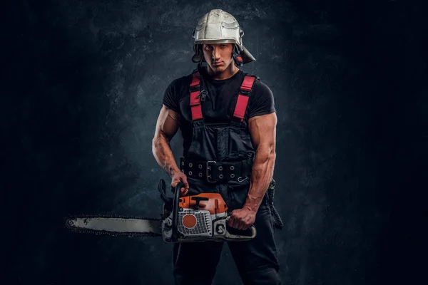 Young lumberjack wearing protective clothes posing with a chainsaw in a dark studio — Stock Photo, Image