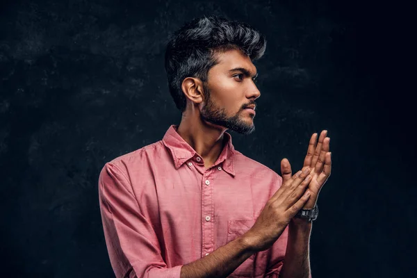 Jovem cara indiano bonito em uma camisa rosa elegante esfrega as mãos e olha para o lado . — Fotografia de Stock