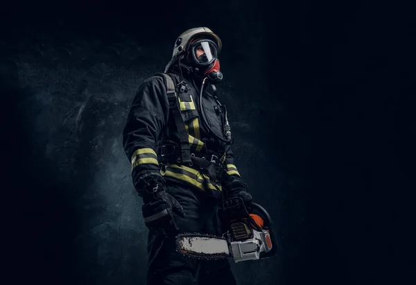 Retrato de um bombeiro em capacete de segurança e máscara de oxigênio segurando uma motosserra. Foto do estúdio contra uma parede de textura escura — Fotografia de Stock