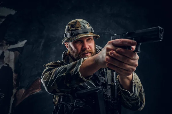 Brutal man in the military camouflaged uniform holding a gun and aiming at the enemy. — Stock Photo, Image