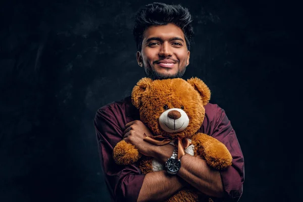 Glimlachend Indiase jongeman in stijlvol shirt hugs teddybeer en zijwaarts op zoek. — Stockfoto