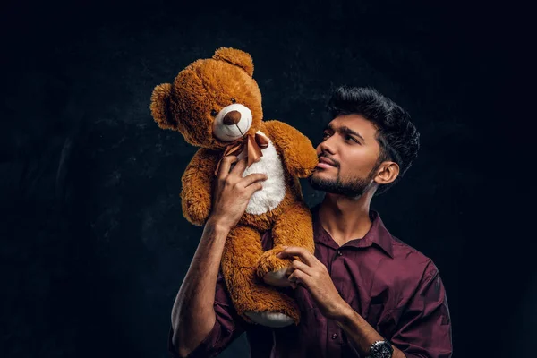 Indiano cara na camisa elegante olha para o seu lindo ursinho de pelúcia, segurando-o nas mãos. Foto do estúdio contra uma parede de textura escura — Fotografia de Stock