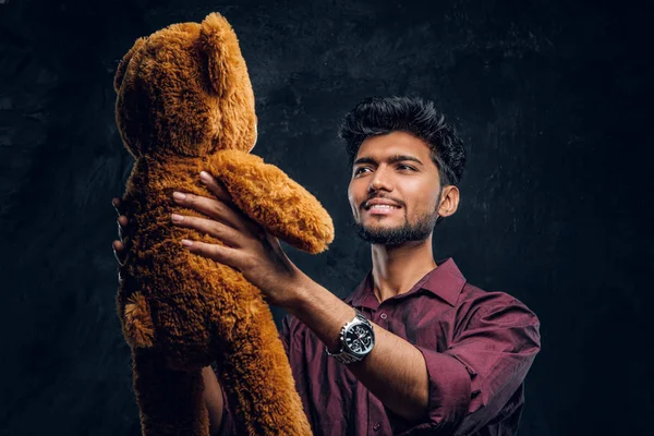 Indiano cara na camisa elegante olha para o seu lindo ursinho de pelúcia, segurando-o nas mãos. Foto do estúdio contra uma parede de textura escura — Fotografia de Stock