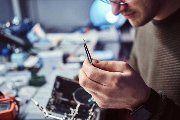 Técnico eletrônico consertando um telefone quebrado, olhando atentamente para o pequeno parafuso segurando-o com pinças — Fotografia de Stock