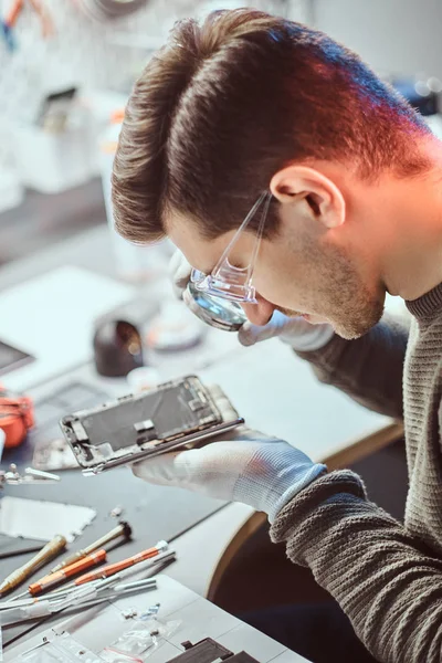 O técnico usa uma lupa para inspecionar cuidadosamente as partes internas do smartphone em uma oficina de reparo moderna — Fotografia de Stock