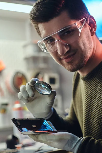 Technicien en lunettes tenant un smartphone endommagé et une loupe, regardant une caméra dans un atelier de réparation moderne — Photo