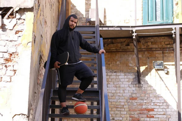 Afro-amerikansk kille i en svart hoodie står på stål trappa håller på en skateboard och tittar på en kamera utomhus — Stockfoto