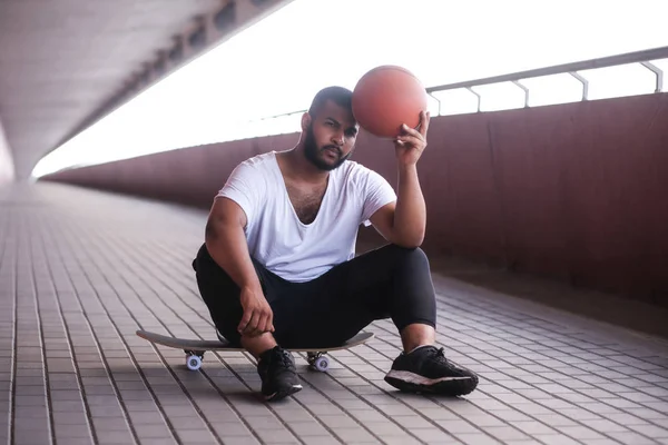 Afroamerikansk man sitter på en skateboard och hålla en basketboll på en bro trottoar — Stockfoto