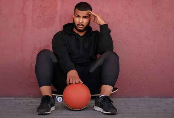 Hombre afroamericano con una sudadera con capucha negra sentado en un monopatín y sosteniendo una pelota de baloncesto mientras se apoya en una pared afuera —  Fotos de Stock