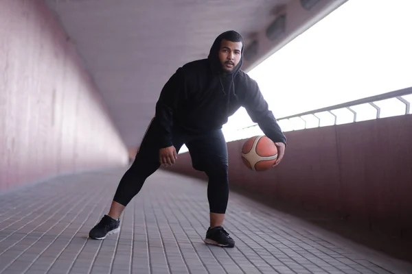 Afro-Amerikaanse man, het dragen van een zwarte hoodie spelen basketbal op de stoep van een brug — Stockfoto