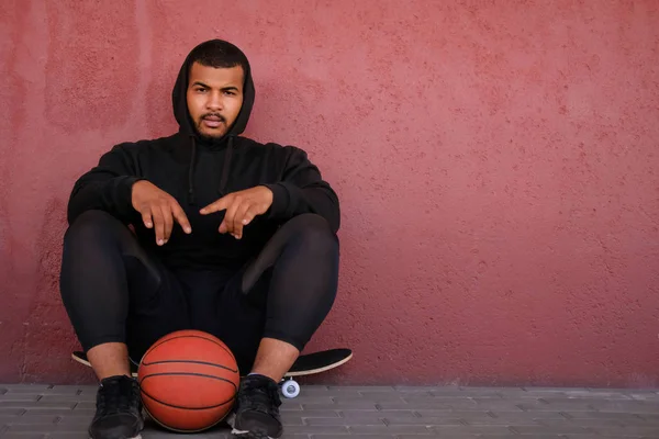 Afro-amerikansk kille klädd i en svart hoodie sitter på en skateboard och hålla en basket medan lutande på en vägg utanför — Stockfoto