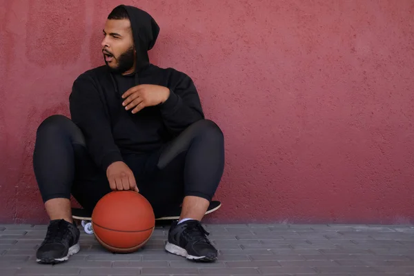 Afroamerikansk man klädd i en svart hoodie som sitter på en skateboard med en basketboll, gäspande och tittar i sidled — Stockfoto