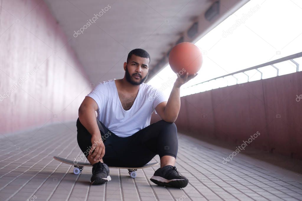 African-American man is sitting on a skateboard and holding a basketball on a bridge sidewalk
