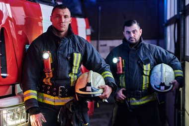 İki itfaiyeci itfaiye kamyonu yanında koruyucu Tekdüzen durduğunu bir itfaiye garajda giyiyor.