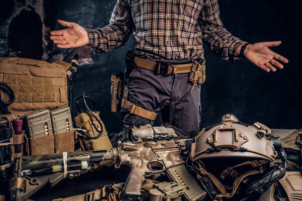 Man in a checkered shirt showing his military uniform and equipment. Modern special forces equipment. Studio photo against a dark textured wall