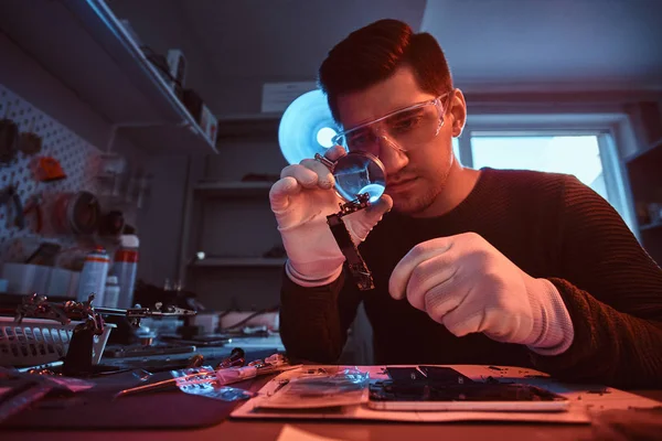 Electronic technician mending a broken tablet carefully examines the chip of the tablet using a magnifying glass. Illumination with red and blue lights — Stock Photo, Image