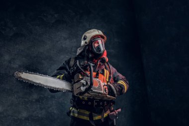 Kask ve oksijen maskesi bir chainsaw tutan bir itfaiyeci portresi. Studio fotoğraf bir karanlık dokulu duvara