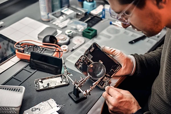 Servicemann repariert beschädigtes Smartphone in der Werkstatt mit Lupe und Pinzette. — Stockfoto