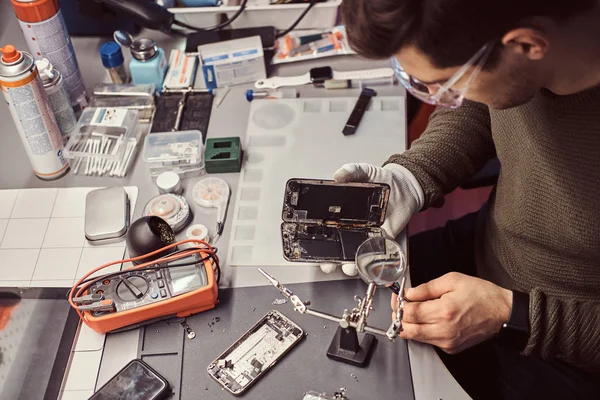 Servicemann repariert beschädigtes Smartphone in der Werkstatt mit Lupe und Pinzette. — Stockfoto