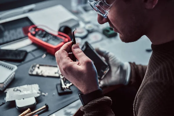 Técnico eletrônico consertando um telefone quebrado, olhando atentamente para o pequeno parafuso segurando-o com pinças — Fotografia de Stock