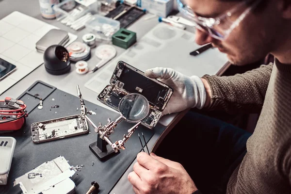Служащий использует увеличитель и пинцет для ремонта поврежденного смартфона в мастерской . — стоковое фото