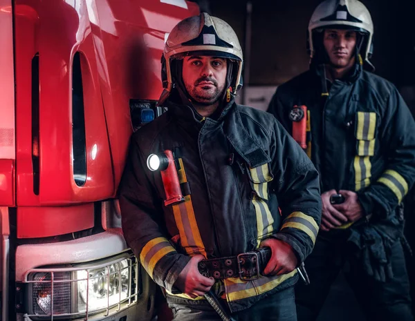 Két tűzoltók visel védő egységes mellette tüzet egy teherautó, egy garázs, egy tűzoltóság. — Stock Fotó