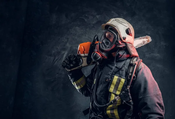 Brandweerman dragen van volledige beschermende uitrusting poseren met een kettingzaag op zijn schouder. Studio foto tegen een donkere getextureerde muur — Stockfoto