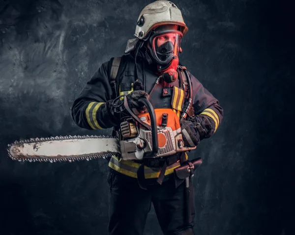 Retrato de um bombeiro em capacete de segurança e máscara de oxigênio segurando uma motosserra. Foto do estúdio contra uma parede de textura escura — Fotografia de Stock