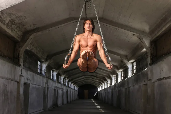 Un deportista profesional con un hermoso cuerpo muscular entrena en anillos gimnásticos en un edificio industrial abandonado —  Fotos de Stock